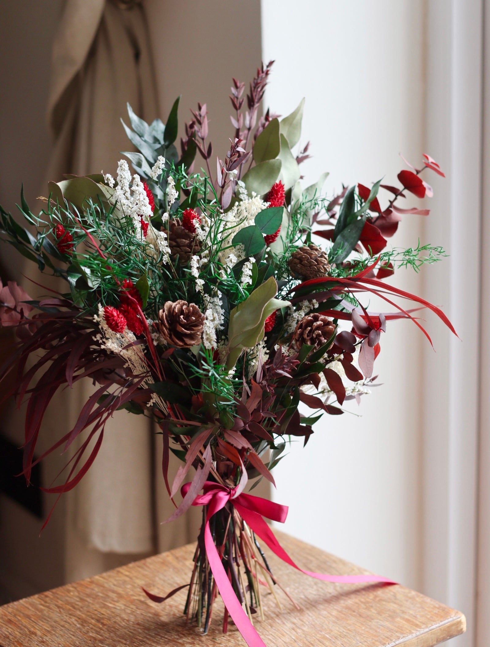 christmas dried flower bouquet in dark green and red eucalyptus, pine cones and red and white dried flower bouquet sitting on a rustic stool
