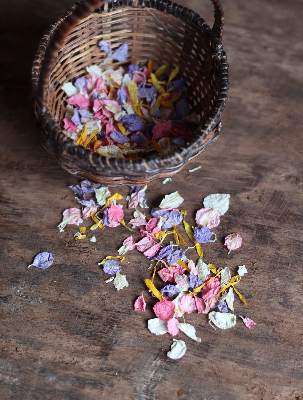 A small woven basket filled with colorful dried flower petals spilled onto a rustic wooden surface.