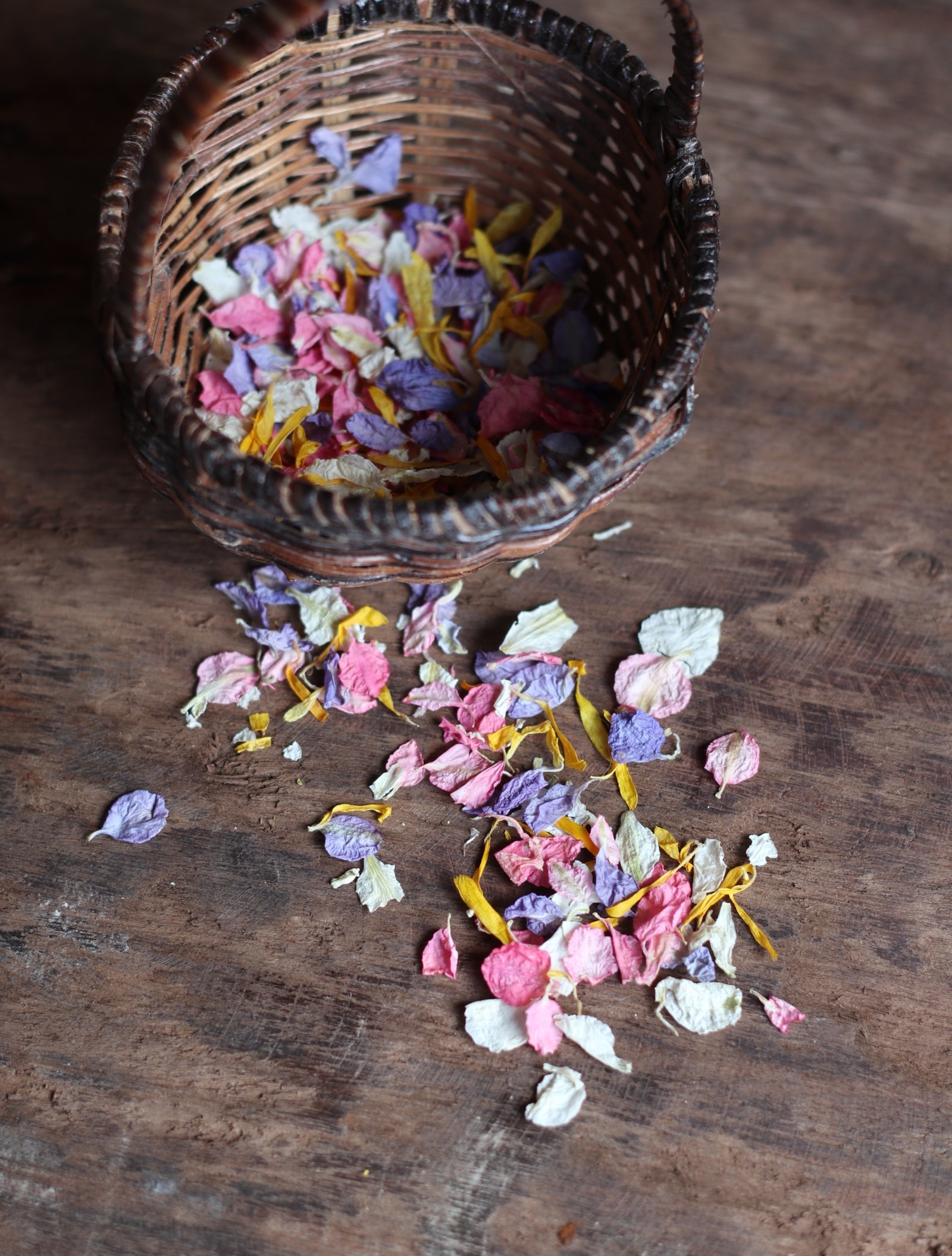 A small woven basket filled with colorful dried flower petals spilled onto a rustic wooden surface.