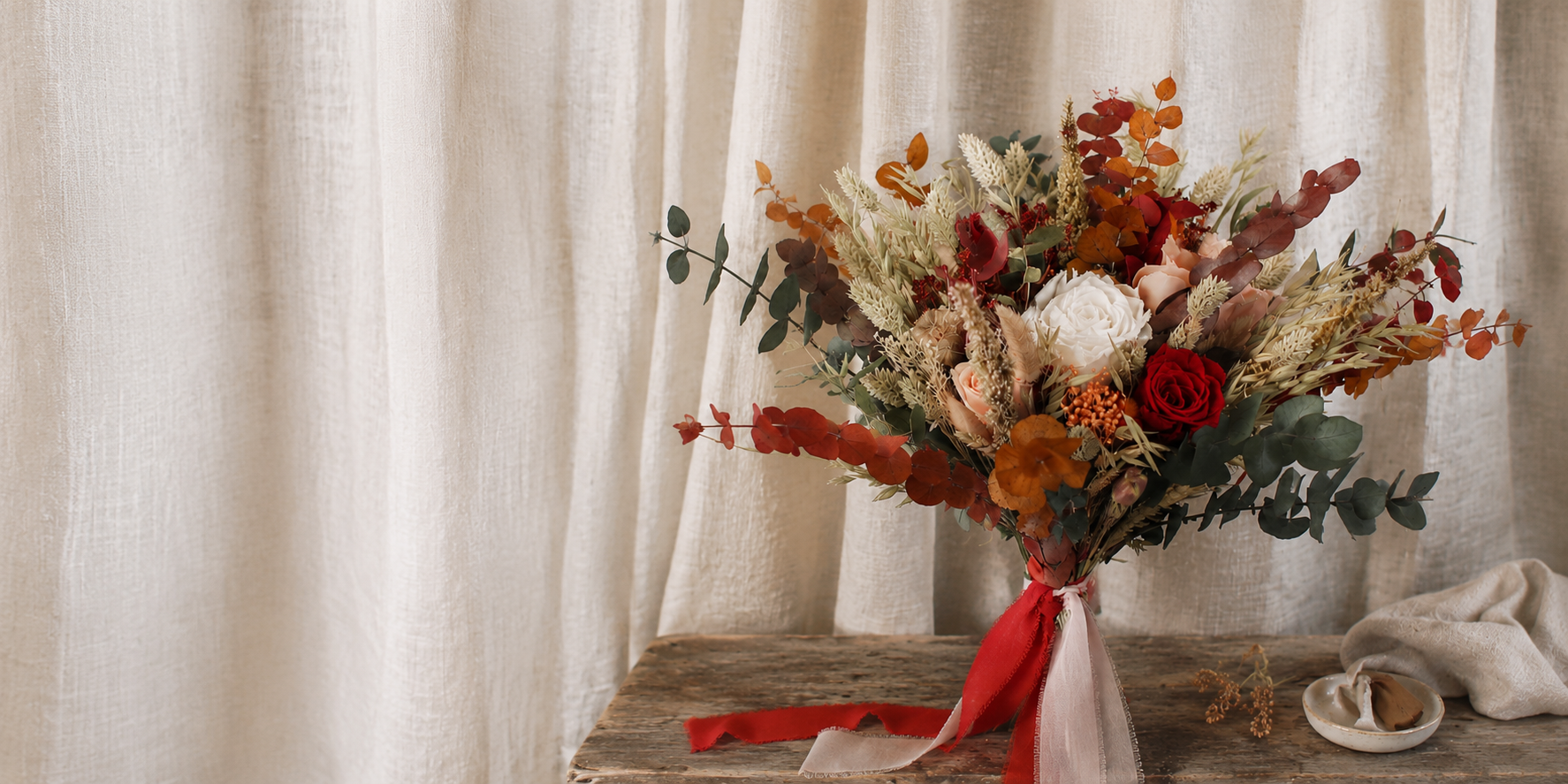 autumn wedding flower bouquet in oranges, peaches, sage greens and burgundy, on a rustic stool with added chiffon ribbons