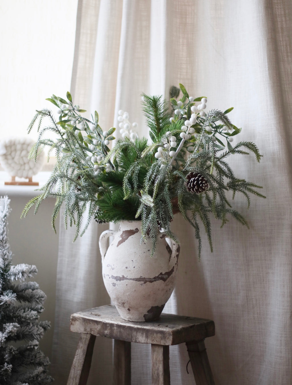 Decorative arrangement of greenery and berries in a textured vase on a wooden stool against a white curtain.