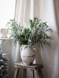Decorative arrangement of greenery and berries in a textured vase on a wooden stool against a white curtain.