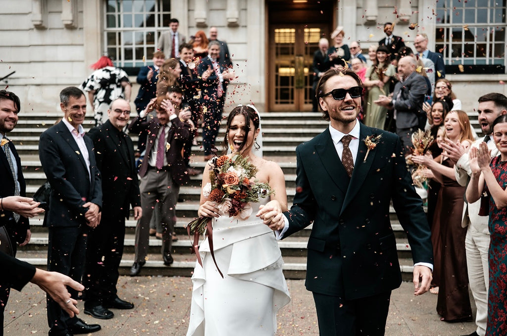 Wedding couple walking down steps  holding a dried flower bouquet with guests clapping, wearing sunglasses.