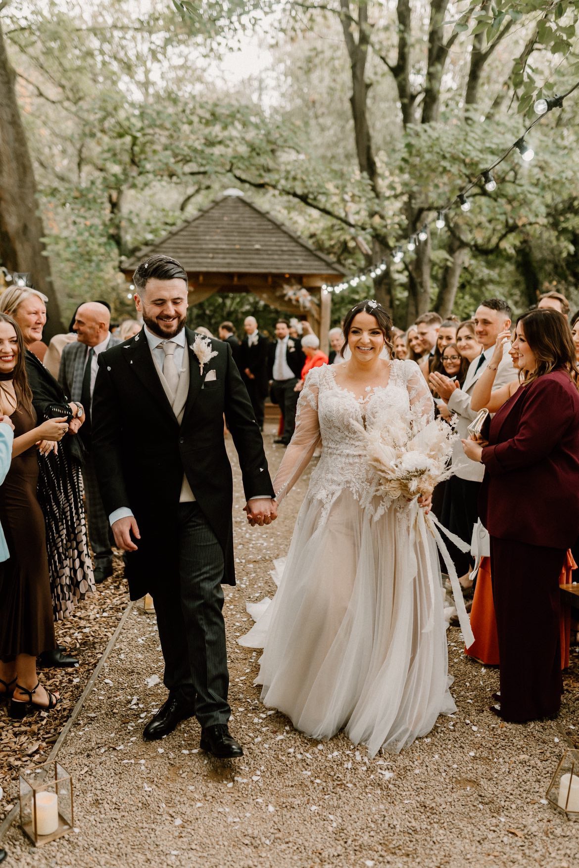 Wedding couple walking down a path with guests on either side holding a dried flower bouquet